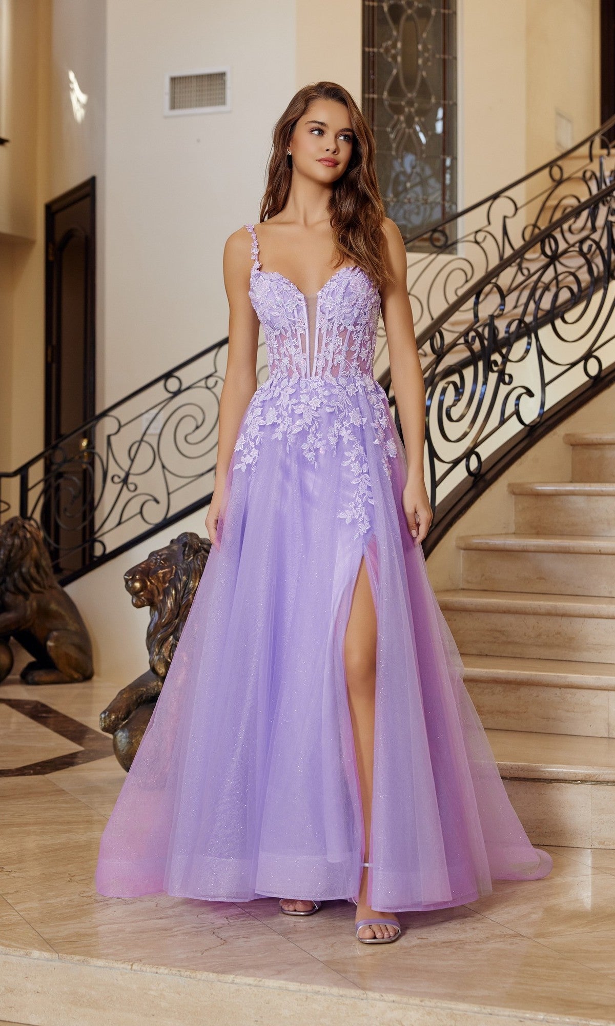A woman stands indoors on a staircase, wearing the Nox Anabel R1824 long prom dress in lilac, featuring a sleeveless design, floral appliqué and embroidery, corset bodice, and a thigh-high slit.